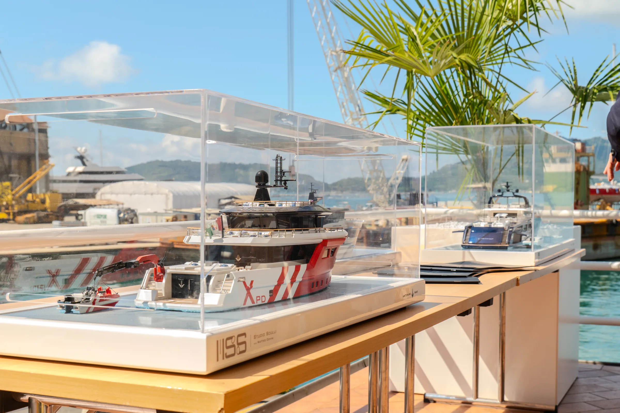 Modelli di navi in teche di vetro esposti in banchina al cantiere Antonini Navi, con lo scafo di uno yacht sullo sfondo