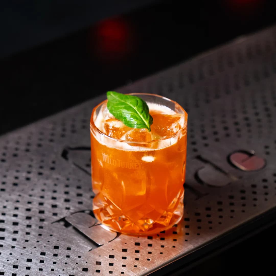 Cocktail arancione con foglia di basilico appoggiato sul bancone metallico