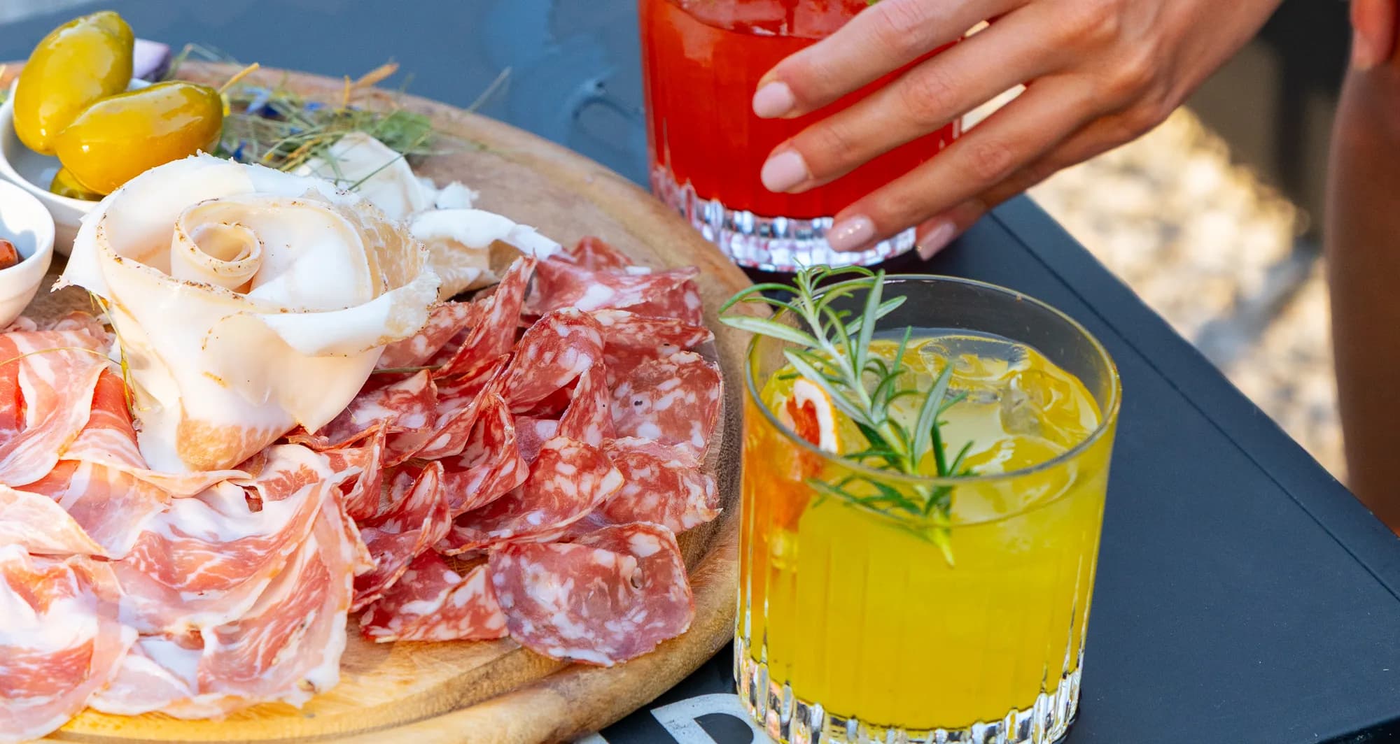 Tagliere di salumi e formaggi con due cocktail sul tavolo del PiN, una mano che prende il bicchiere