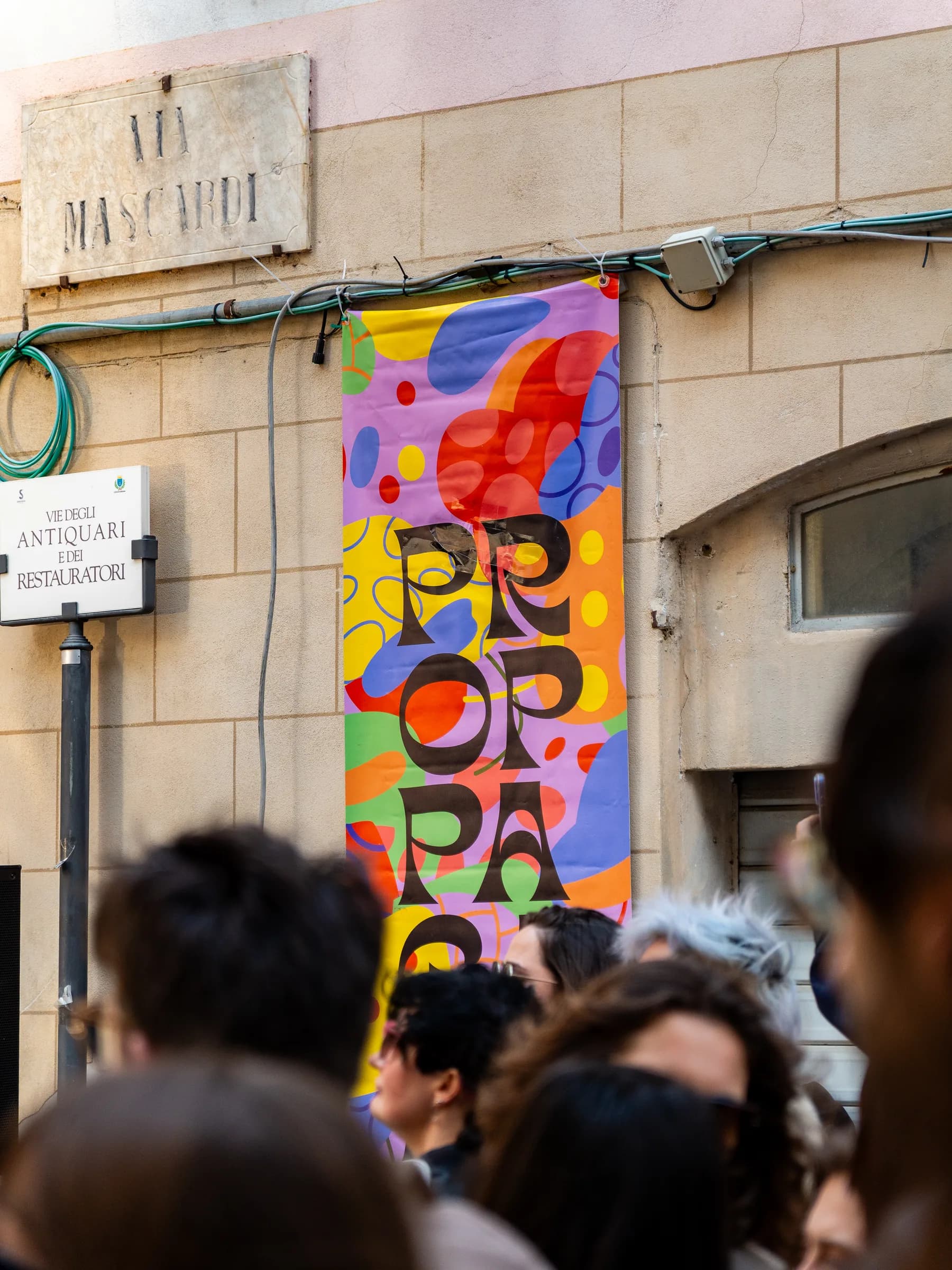 Manifesto colorato del festival Propaganda appeso al muro di un palazzo, con il pubblico sotto