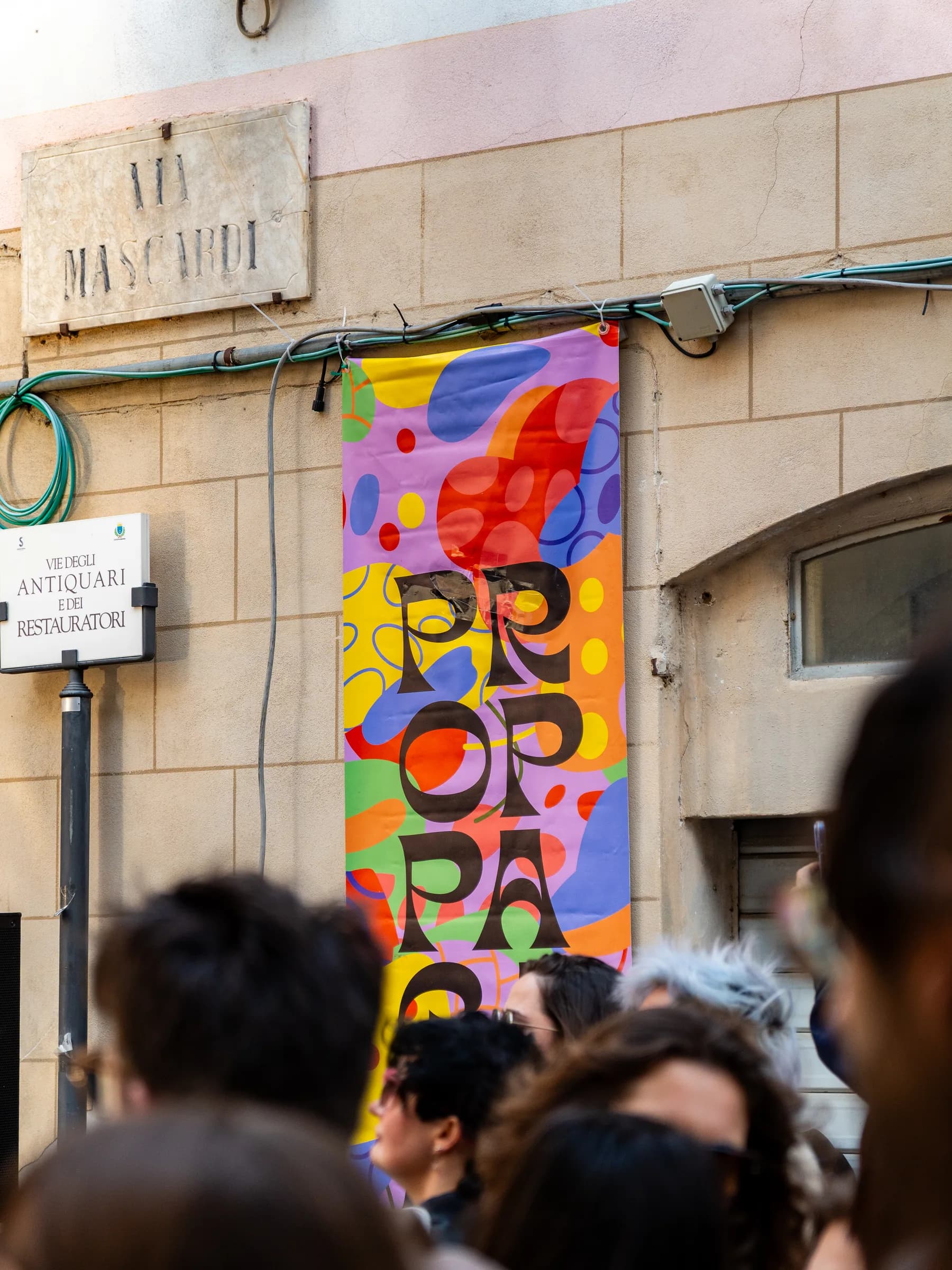 Manifesto colorato del festival Propaganda appeso al muro di un palazzo, con il pubblico sotto