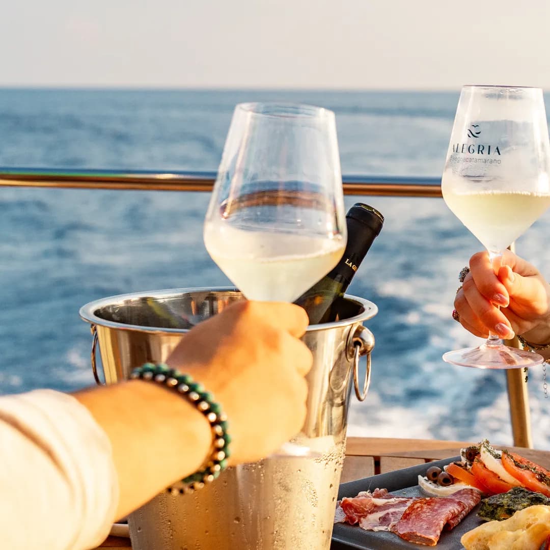 Brindisi con due calici di vino bianco sopra un tagliere di salumi, a bordo dell'Alegria